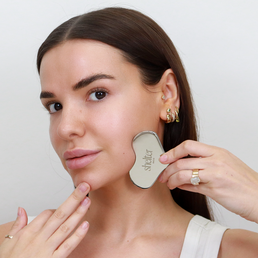 Contouring Stainless Steel Gua Sha held against the model's face, showcasing its sleek design and reflective surface.