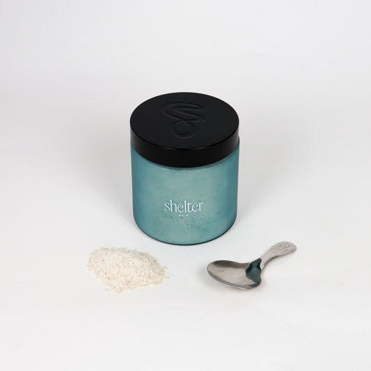 Coconut Milk Bath Melt in a frosted blue jar with a black lid, with white bath melt powder next to a metal spoon. 