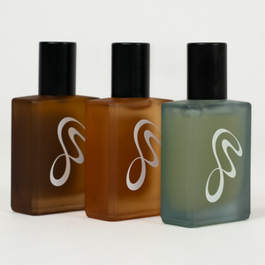 Three frosted glass perfume bottles with black caps arranged in a row on a light gray background. The bottles contain amber-brown liquid in the center and a muted sage-green liquid on the right, with a white stylized logo on each bottle.