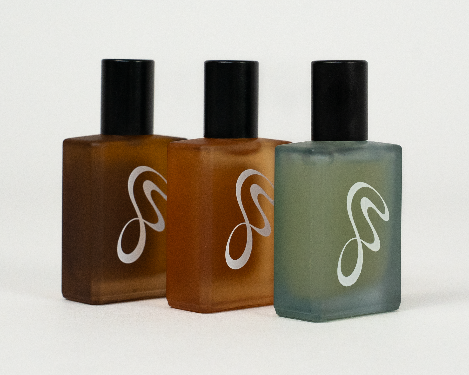 Three frosted glass perfume bottles with black caps arranged in a row on a light gray background. The bottles contain amber-brown liquid in the center and a muted sage-green liquid on the right, with a white stylized logo on each bottle.